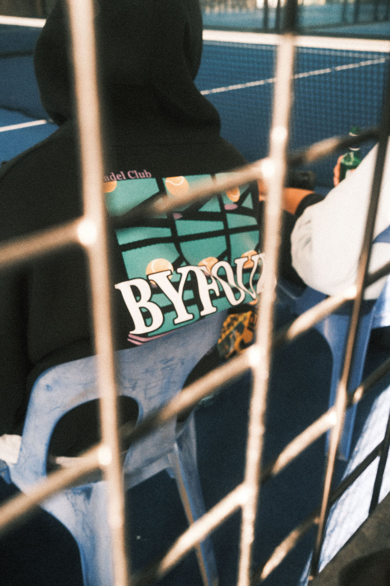 close up through a fence of a padel players back and the byfour hoodie backprint
